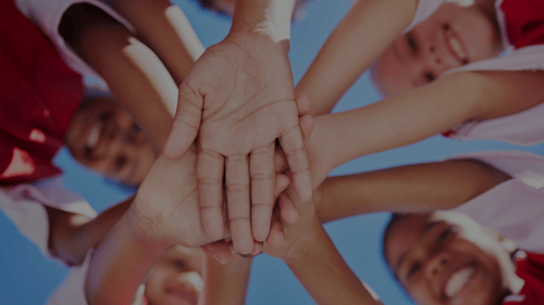 Camera pointing up at elementary aged kids in a circle, looking down, with their hands stacked on top of each other and ready to throw in the air in celebration.