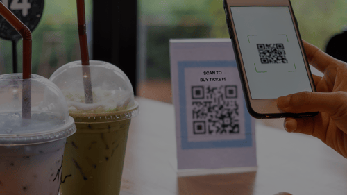 Hand holding a phone and scanning a small plastic stand with a poster in it, with a QR code on the poster. Two iced coffees, one green matcha and one a pink cream colour are beside the poster.