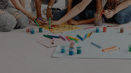 Four girls sitting down, with only their hands and legs showing, drawing with paints and colored pencils on paper on the floor.