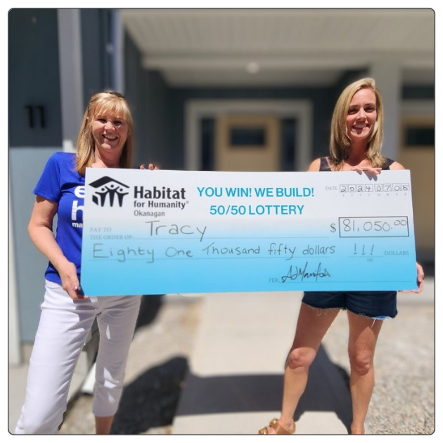 Two individuals smile at the camera while holding a large cheque from both sides, as the CEO of Habitat for Humanity Okanagan presents the latest raffle winner with their winnings