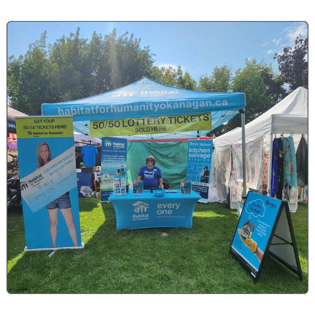 Photo of an outdoor fundraising booth on a sunny day, featuring Habitat for Humanity Okanagan's raffle branding. A happy volunteer stands at the table, ready to chat and sell raffle tickets to event attendees