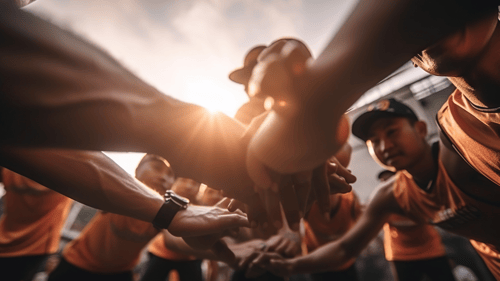 Group of youth in sports jerseys and ball caps holding hands in a circle, with the sun shining through their fingers